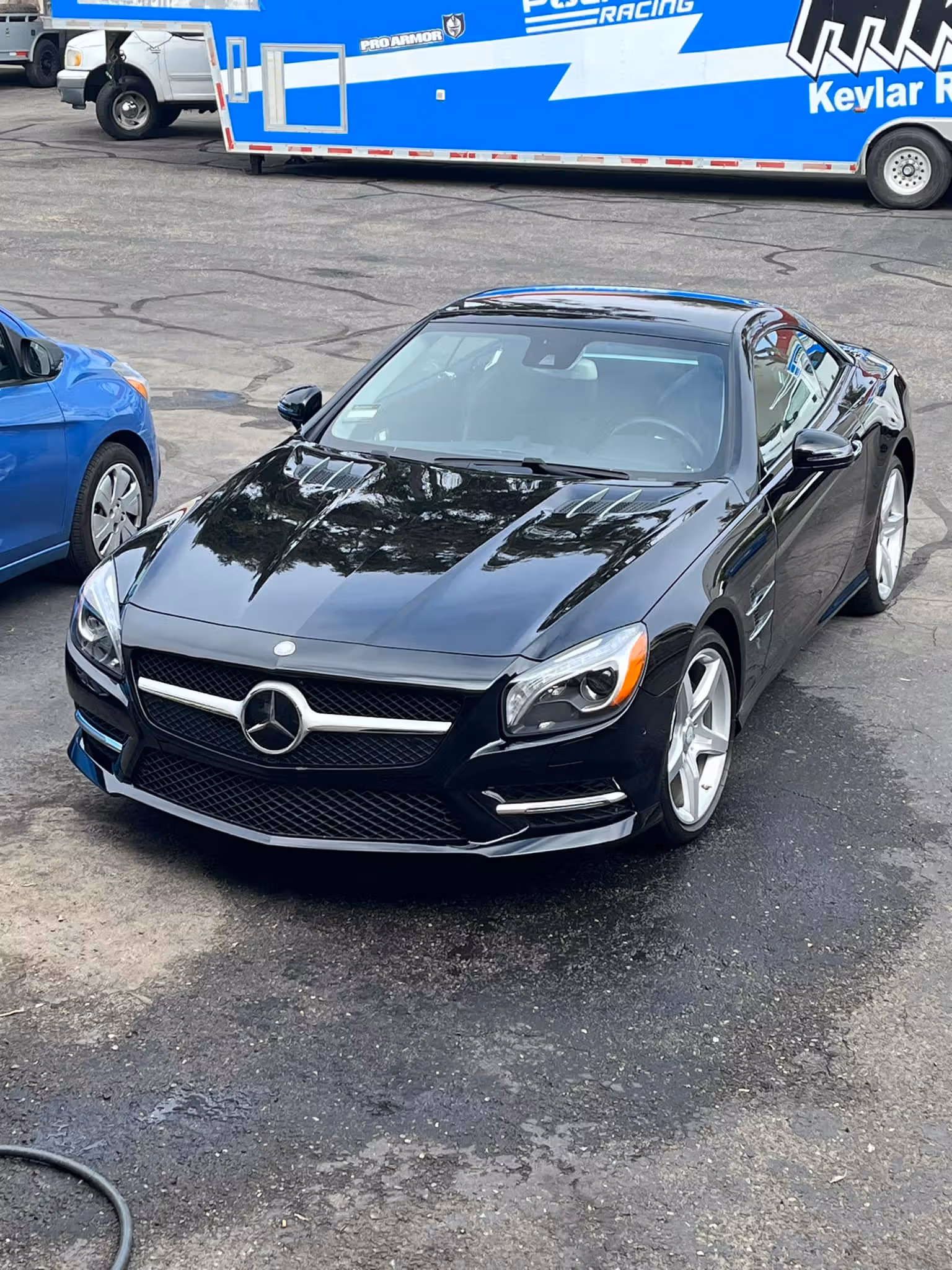 Black Mercedes-Benz sports car parked on asphalt with a blue car and trailer in background.