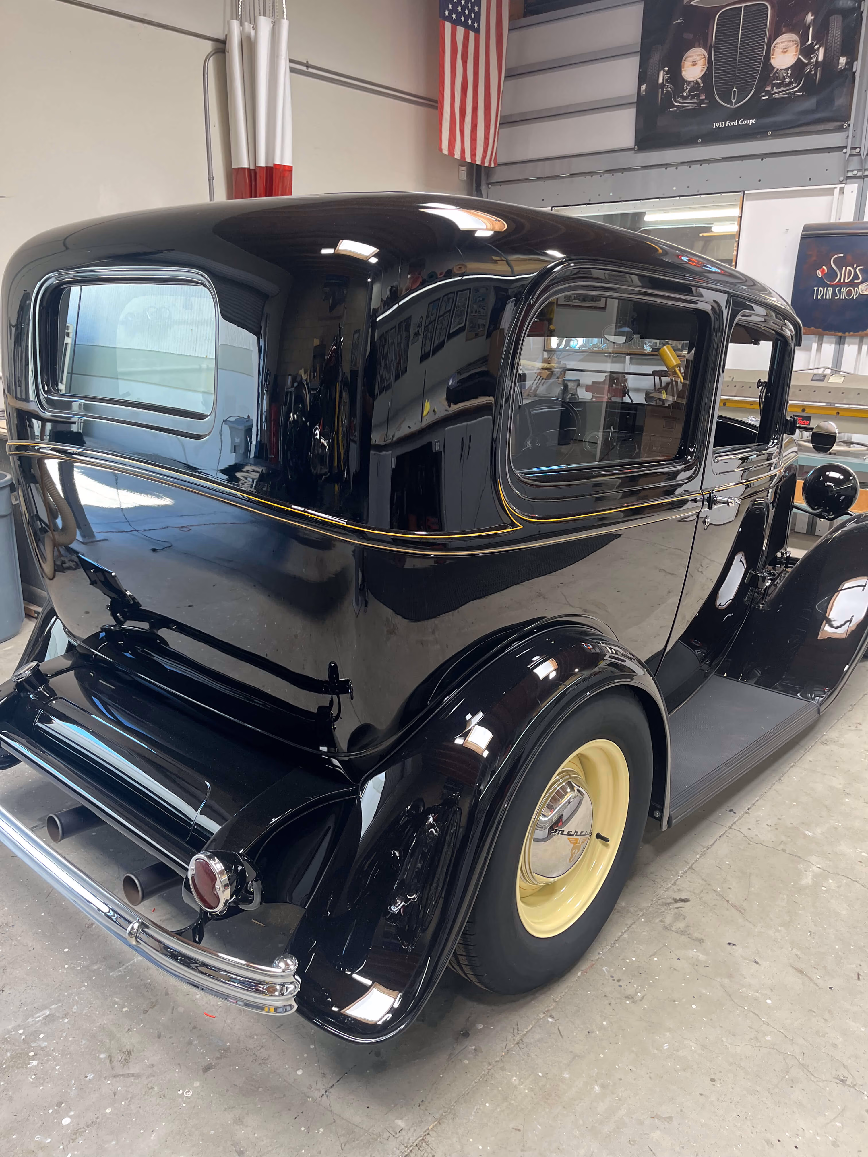 Black vintage car with yellow wheels parked in a garage with an American flag on the wall.