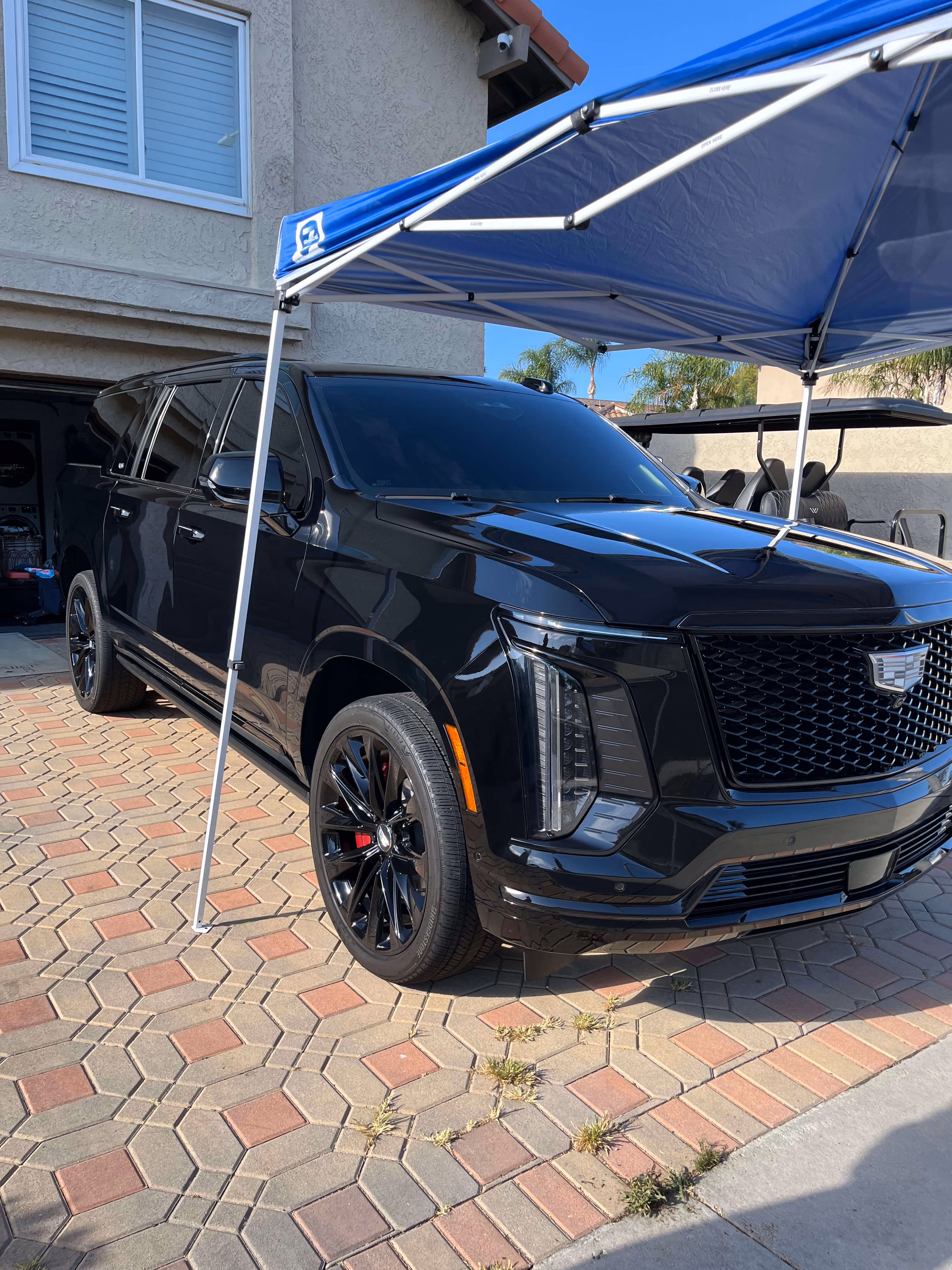 Black Cadillac SUV parked on patterned driveway under a blue canopy beside a house.