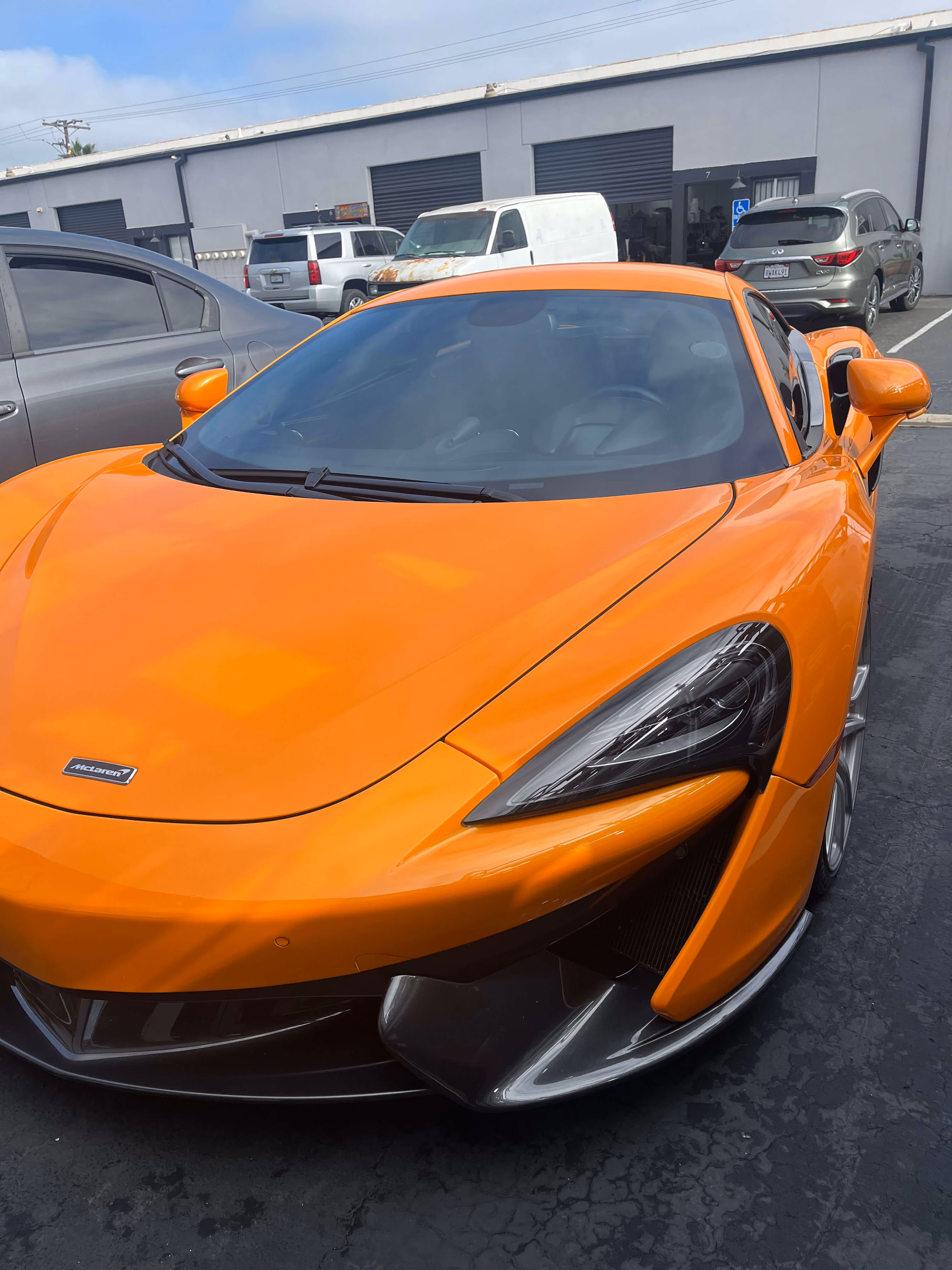 Front view of an orange McLaren sports car parked on asphalt near other vehicles and buildings.