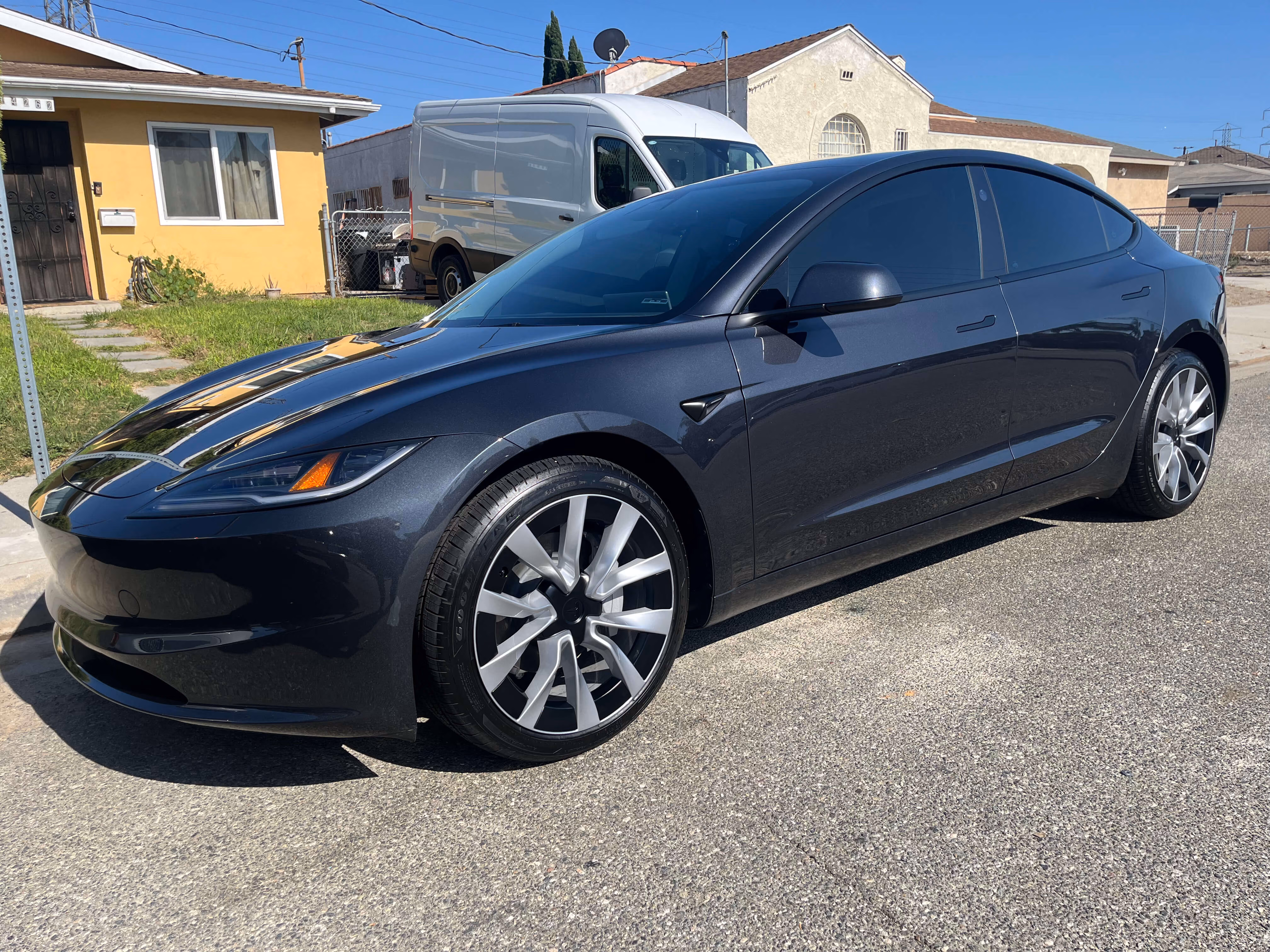 Dark blue Tesla Model 3 parked on a residential street with houses and a white van in the background.