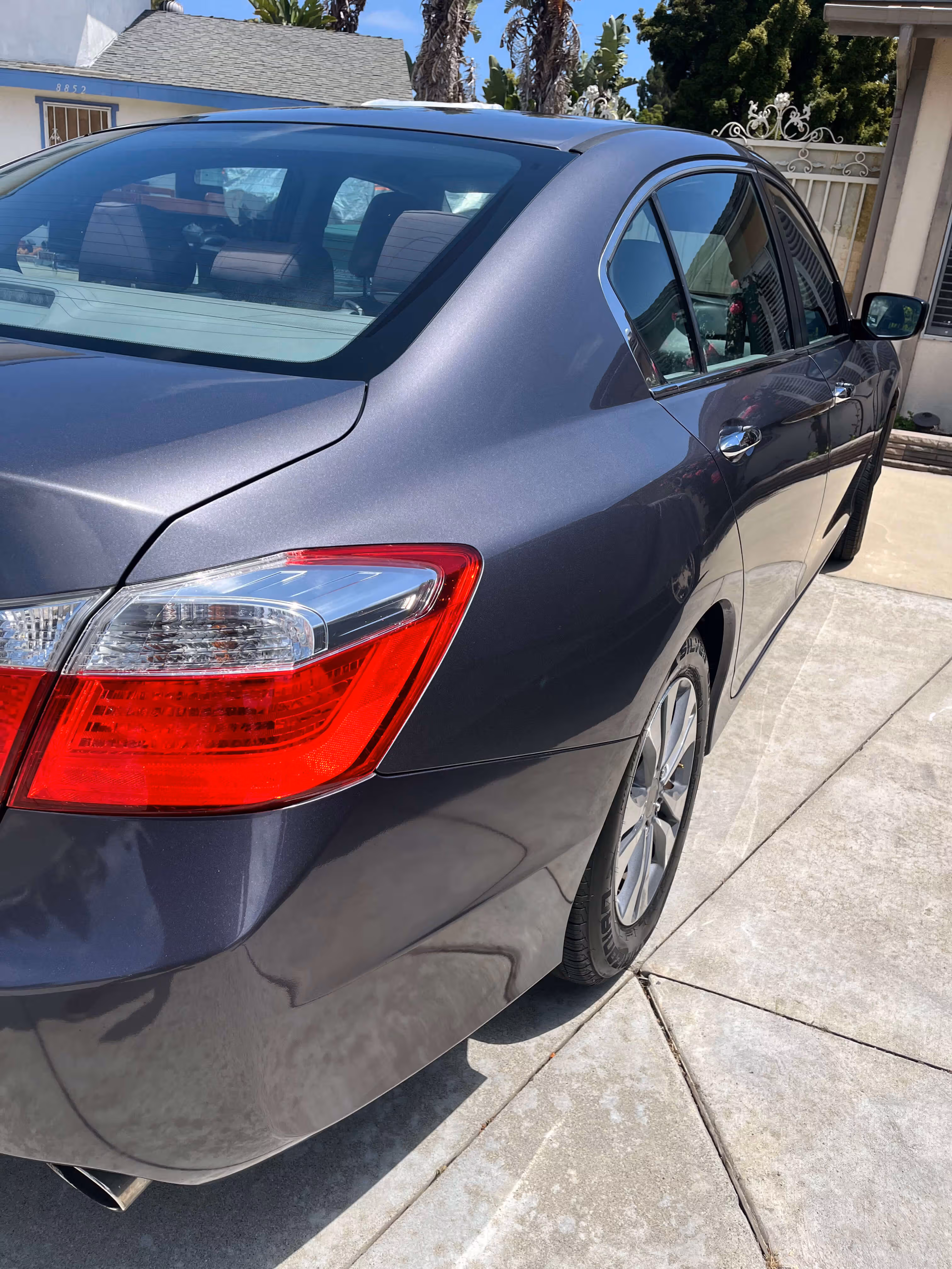 Rear side view of a dark gray sedan car parked on a concrete driveway on a sunny day.
