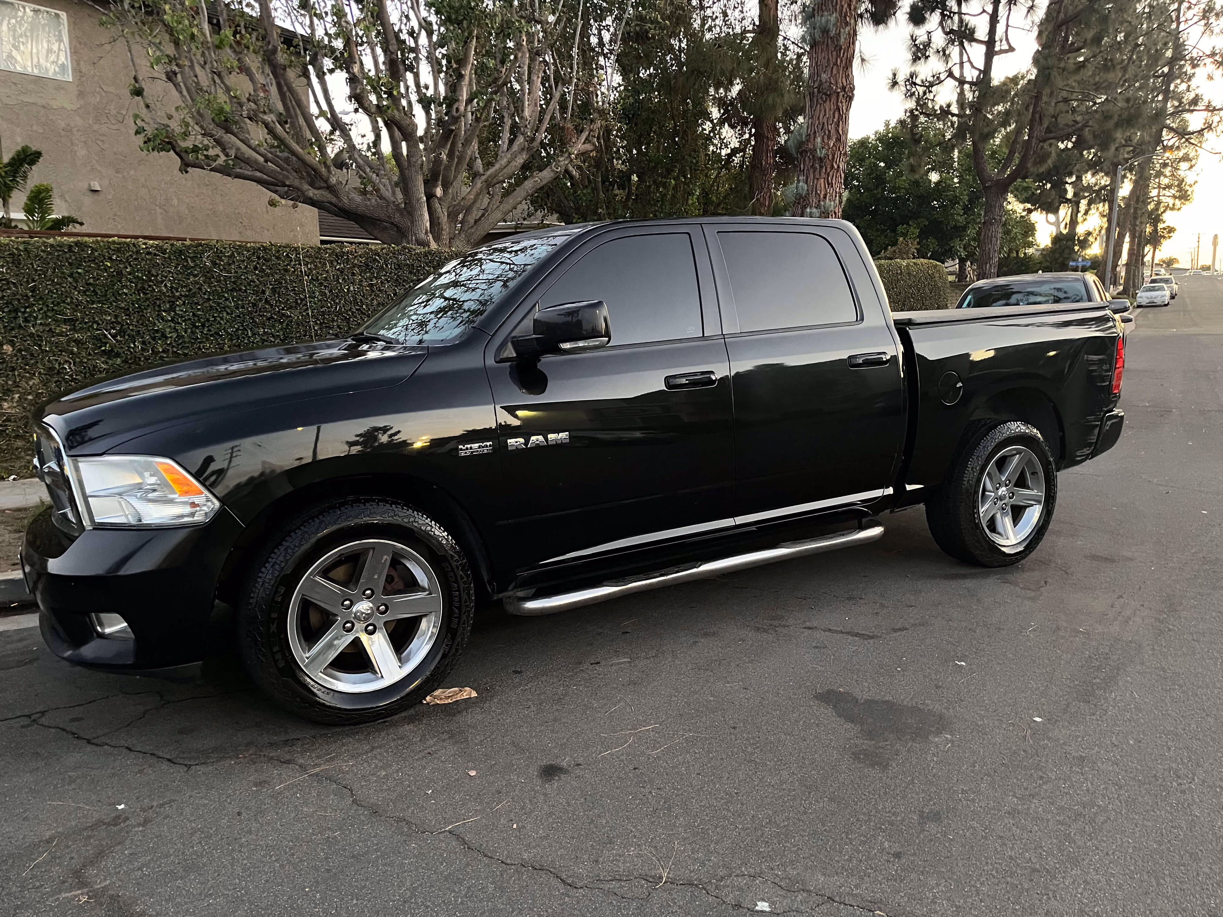 Black Dodge Ram pickup truck parked on a residential street near trees and houses at dusk.
