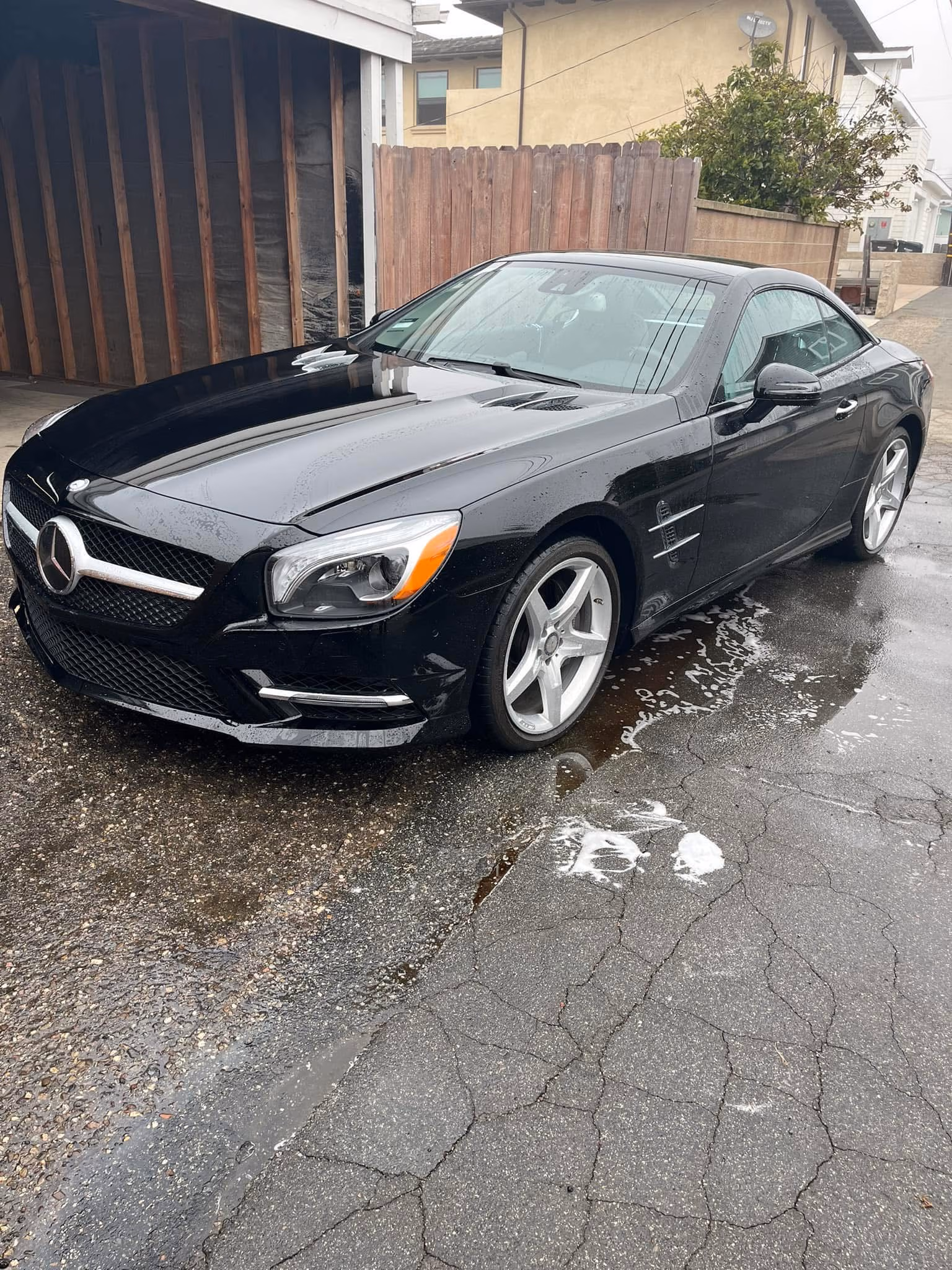Black Mercedes convertible parked on a wet cracked driveway near a wooden fence and house.