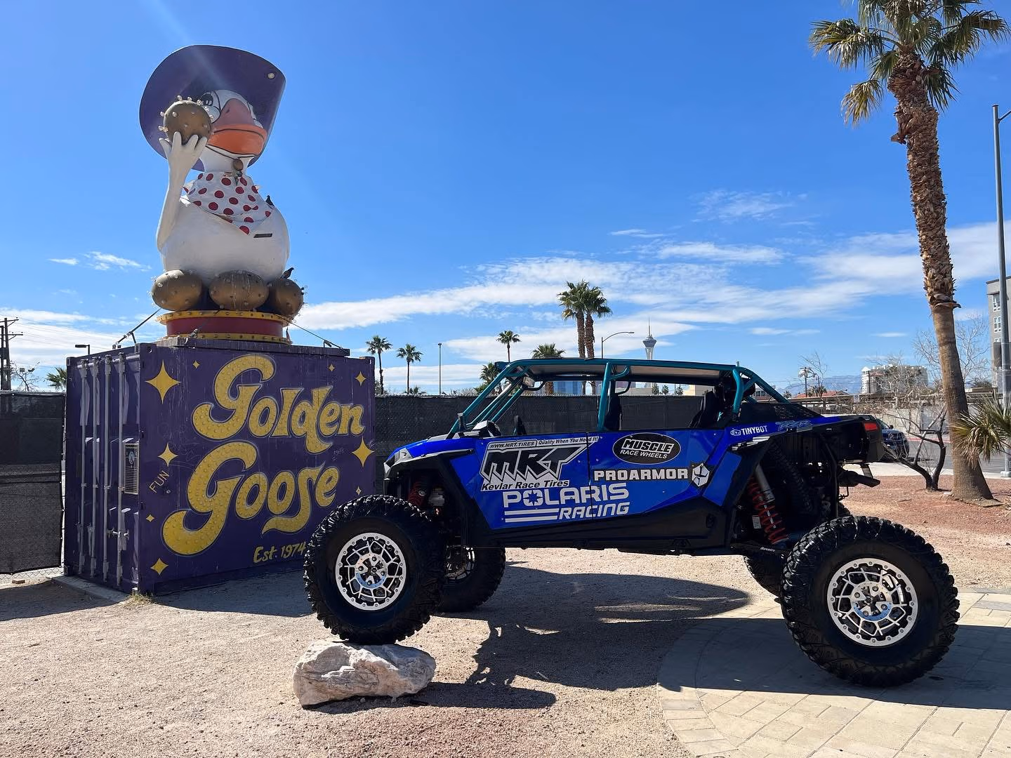 Blue Polaris Racing off-road vehicle next to a purple Golden Goose container with large goose statue on top.