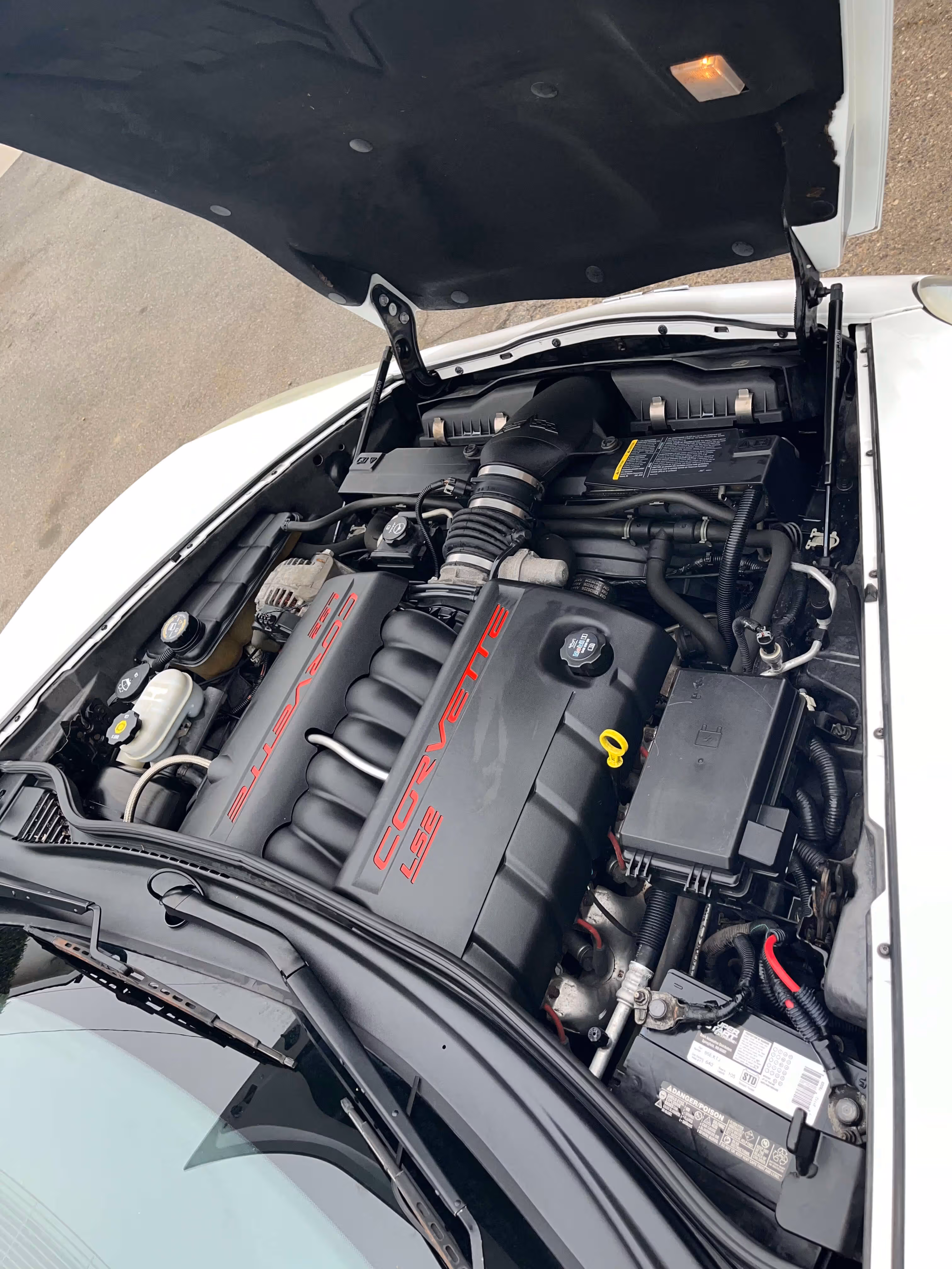 Open hood of a white car revealing a Corvette LS2 engine with visible battery and fluid reservoirs.