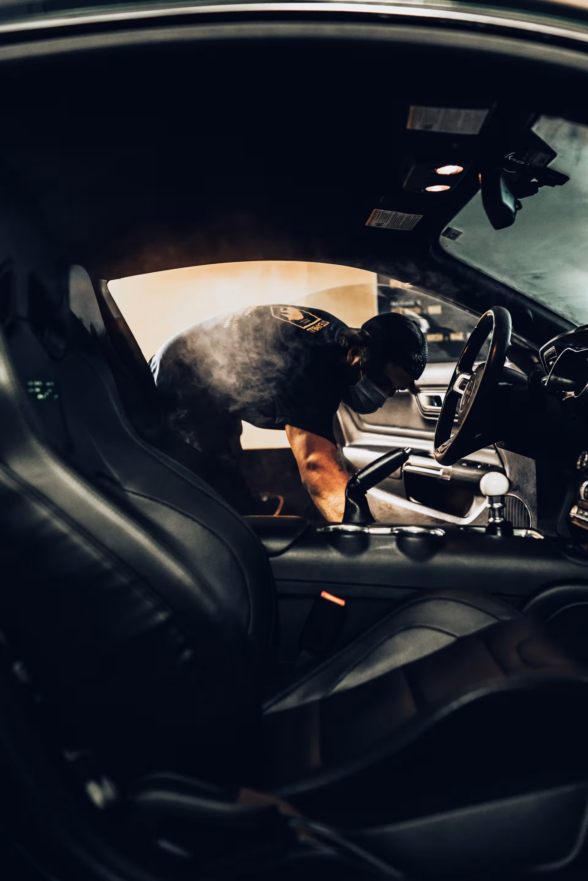 Person wearing mask steam cleaning the interior of a black car, with steam visible inside.